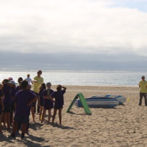 A group of children in purple shirts listens attentively to an instructor on the beach, with surfboards nearby and ocean scenery in the background.