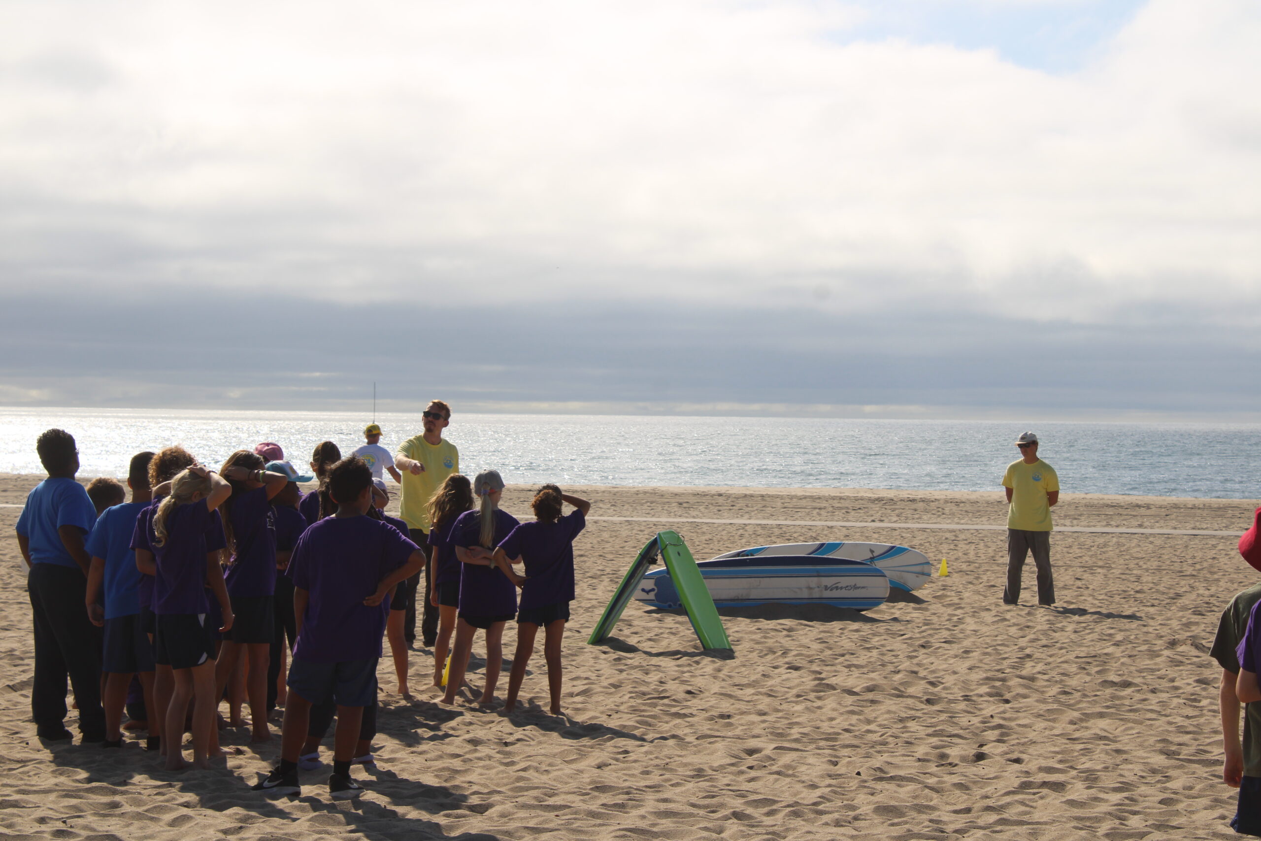 A group of children in purple shirts listens attentively to an instructor on the beach, with surfboards nearby and ocean scenery in the background.