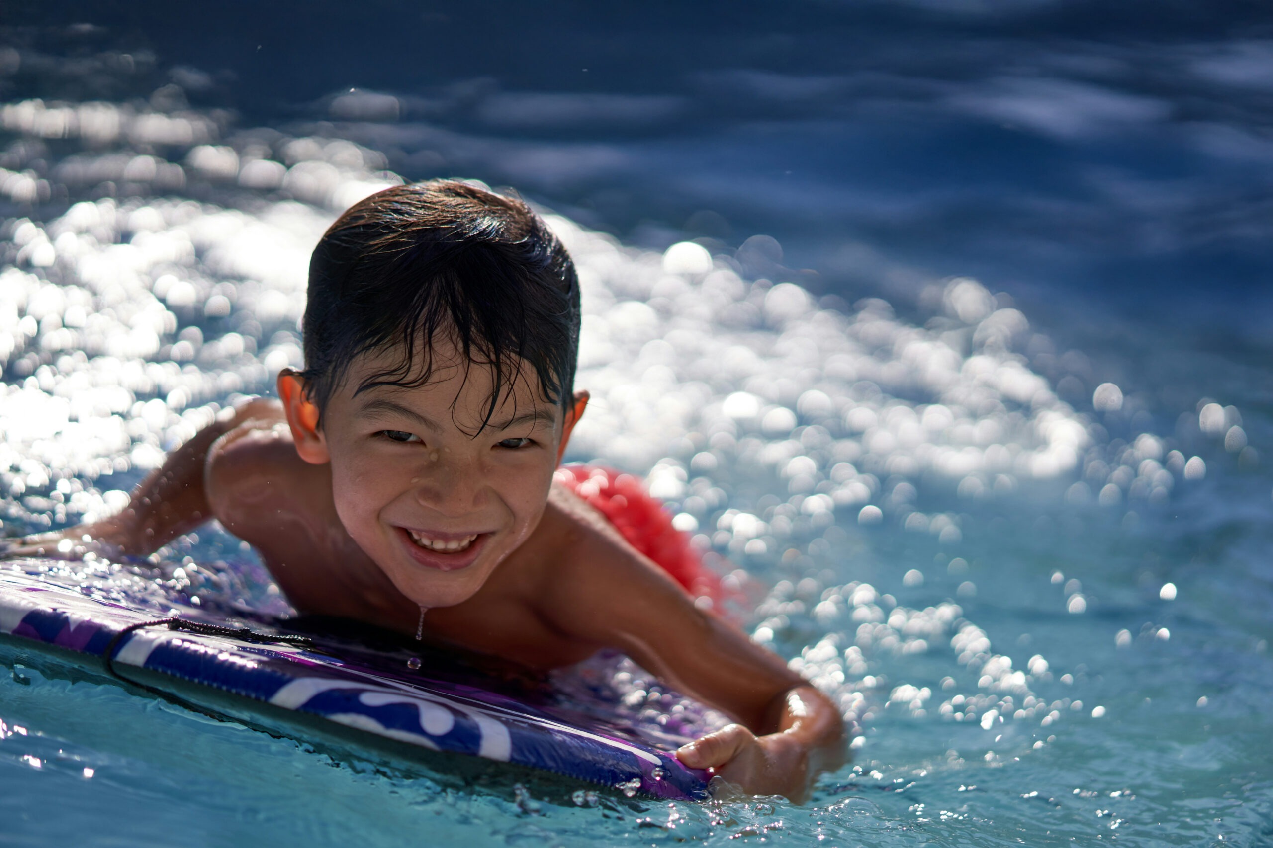 Young boy with a big smile swims in a pool, lying on a boogie board, surrounded by sparkling water droplets. Fun summer activity.