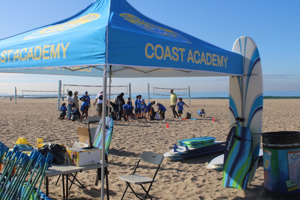 Coast Academy tent at the beach, with children in blue shirts engaging in activities and volleyball courts in the background, promoting outdoor sports.